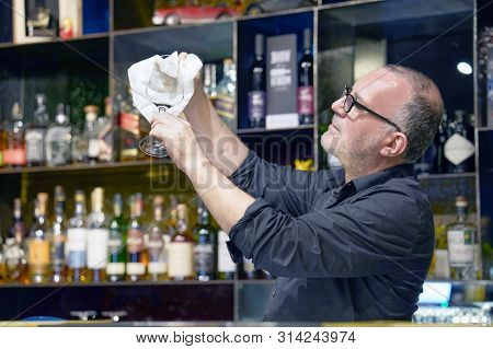 Elderly Bartender Cleans The Glass. A Handsome Bartender Polishes A Glass Of Wine Glasses. The Conce