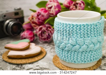 A White Cup Of Tea In A Knitted Blue Case With An Old Camera And Pink Flowers Tulips And Sweets, Gin