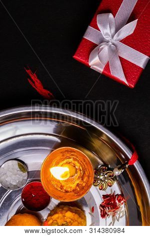 Indian Festival - Elegant Rakhi, Rice Grains, Kumkum, Sweets, Diya In Plate With Red Gift Box On Bla