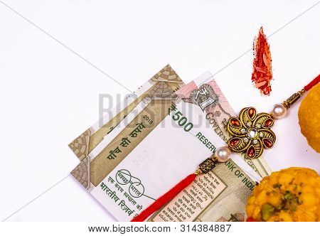 Raksha Bandhan Concept - Elegant Rakhi With Indian Currency, Sweets And Kumkum Rice Grain Mark On Wh