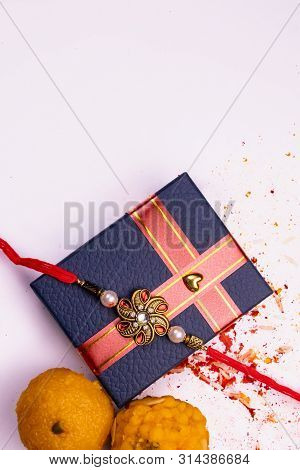 Top View Of Sprinkled Rice, Kumkum, Haldi With Elegant Rakhi On Blue Gift Box And Sweets On White Su