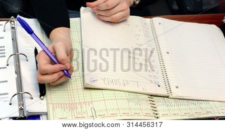 Teacher Grades Papers And Records It In Her Grade Book. She Is Holding A Purple Pen.