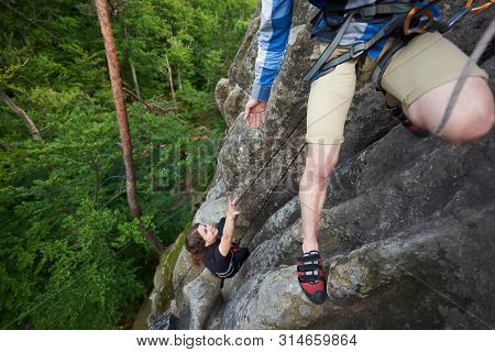 Young Man Rock Climber Climbing With Safety Harness And Rope. Man Hanging On Rock Helping Friend To 