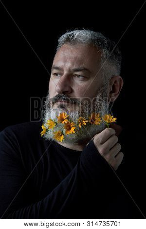Portrait Of A Pensive Brutal Man With A Gray Beard Decorated With Yellow Flowers On A Black Backgrou