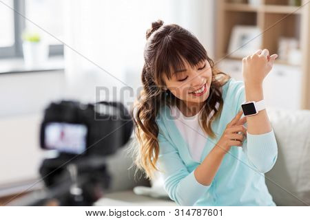 blogging, technology and videoblog concept - happy smiling asian woman or blogger with camera recording video blog of smart watch at home