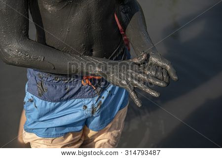 The Body And Arms Of A Teenager, Thickly Smeared With Black Healing Mud, Have Bathing Shorts On It. 