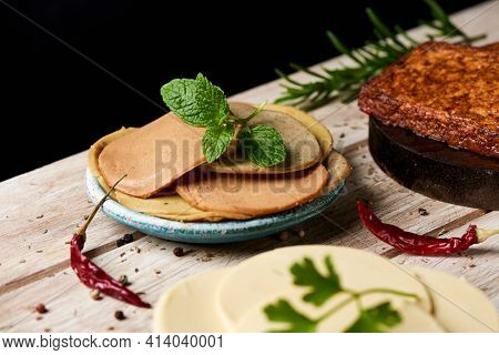 closeup of some some cold cuts made of tofu in a green ceramic plate on a white rustic table, next to some slices of vegan cheese and some tofu fillets