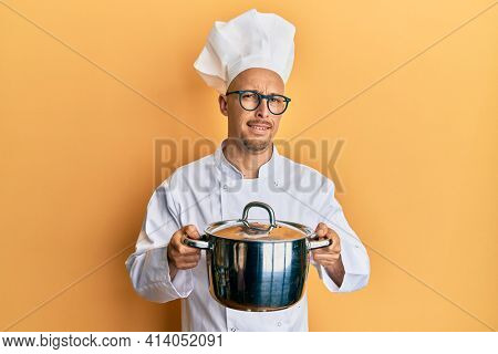 Bald man with beard wearing professional cook holding cooking pot clueless and confused expression. doubt concept. 