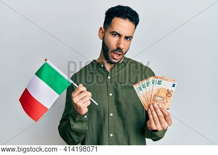 Young hispanic man holding italy flag and euros banknotes in shock face, looking skeptical and sarcastic, surprised with open mouth 