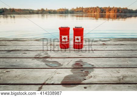Two Cans Of Beer In Red Cozy Beer Can Coolers With Canadian Flag Standing On Wooden Pier By Lake Out