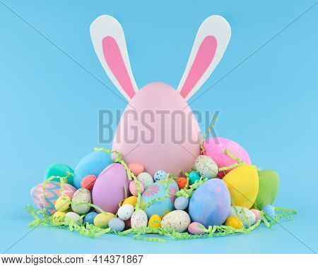 Large pink Easter egg with bunny ears surrounded by a pile of colorful Easter eggs and candy.