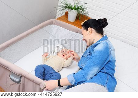 Mother Plays With A Baby Who Is Lying In A Side-bed For A Newborn Or In A Modern Playpen