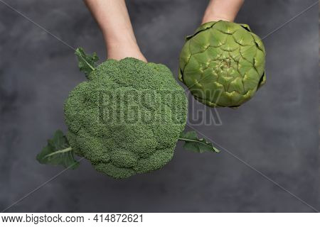 Caucasian Male Hands Holding Brocoli And Artichoke Isolated On Gray Background.