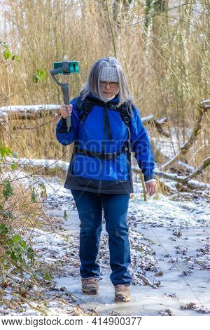 Mature Woman Walking On A Frozen Dirt Trail Surrounded By Wild Vegetation And Snow With Her Mobile P