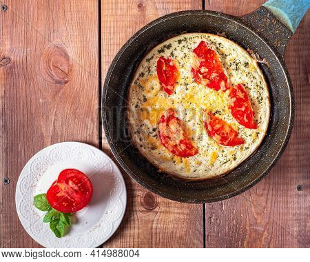 Omelet With Tomatoes And Cheese In A Round Frying Pan And Half A Tomato On A White Plate, On A Woode