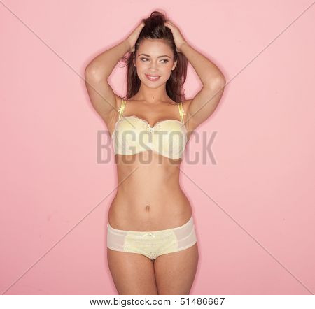 Sexy woman with a lovely body standing posing in yellow lingerie and twiddling her long brunette hair in her fingers, three quarter studio portrait