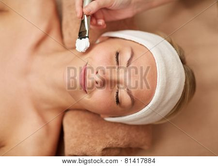 people, beauty, spa, cosmetology and skincare concept - close up of beautiful young woman lying with closed eyes and cosmetologist applying facial mask by brush in spa