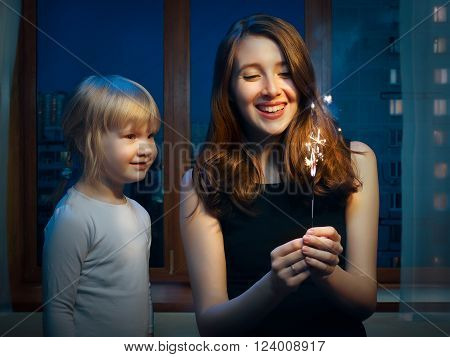An adult woman and a little girl at the window with a sparkler. Girls sister. Outside, the night city. She laughs merrily. The kid looks at the fire sparks