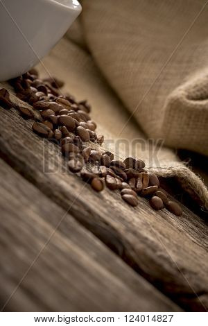Closeup view of coffee beans scattered on rustic textured wooden desk with burlap sac in background and part of white coffee mug on the left side of the image.