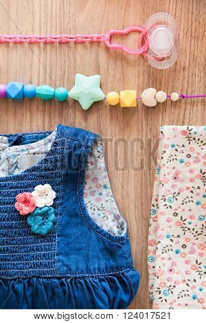 Close up top view of jeans girl's dress decorated with red blue white flovers beige girl's panties with blue brown red flovers pattern and colored silicone beads and pink nipple with pink nipple holder on wooden background