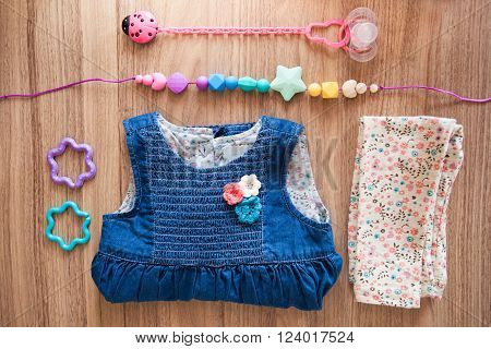 Top view of jeans girl's dress decorated with red blue white flovers beige girl's panties with blue brown red flovers pattern and colored silicone beads and pink nipple with pink ladybird nipple holder and blue and violet plastic stars on wood background