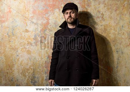 attractive young bearded man in a pea coat and cap is standing at the wall
