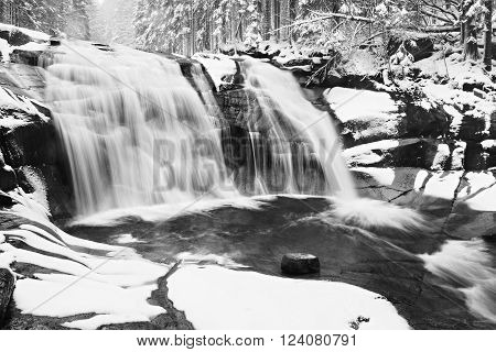 Winter view over snowy boulders to cascade of waterfall. Wavy water level. Stream in deep freeze.