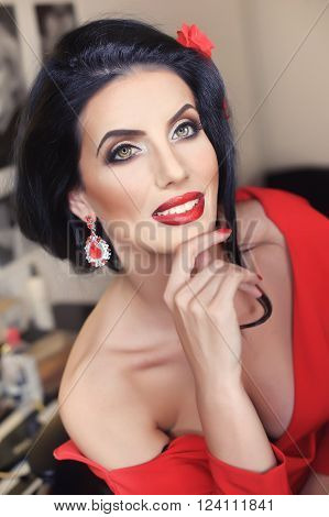 Beautiful young woman with creative make-up and hair style posing. Fashionable attractive brunette with Spanish look, indoors shot. Portrait of lady in red with flower in hair and gorgeous eyes