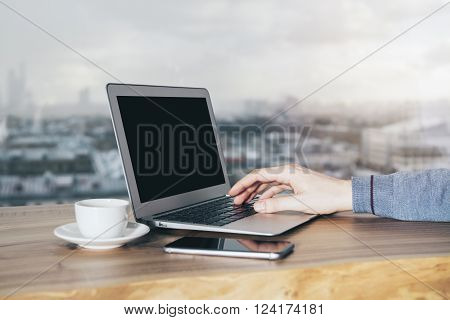 Hand typing on blank laptop keyboard with blurry city in the background. Mock up