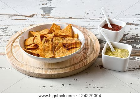Nachos chips in a white bowl over a chopping board with sauces on a wood background