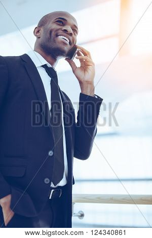 Good budiness talk. Low angle view of cheerful young African man in formalwear talking on the mobile phone and smiling