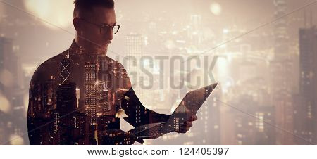 Double exposure bearded businessman wearing black shirt and glasses.Banker holding contemporary notebook hands, online banking.Isolated white, modern skyscraper city at night background