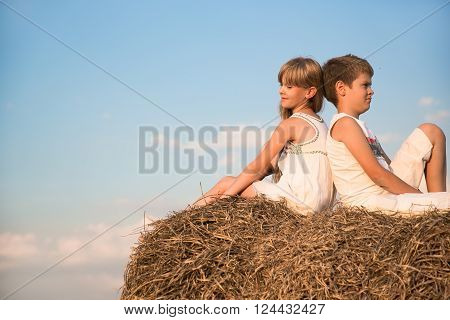 The girl and the boy sit on a haystack backs to each other