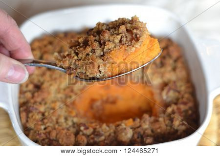 Sweet Potato casserole with crunchy pecan topping; hand holding spoonful of food with casserole dish in background.