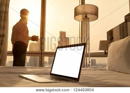 Laptop computer on white bed sheets in cozy room with copy space blank screen. Young businessman with cup of coffee standing at window looking at city scenery on the background