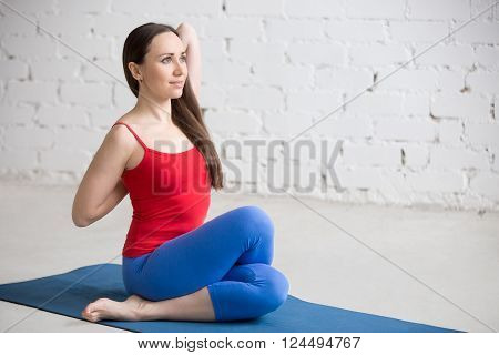 Beautiful happy young woman in bright colorful sportswear working out indoors in white loft interior. Girl sitting in Gomukhasana Cowface posture on blue mat. Yin yoga Shoelace pose. Full length