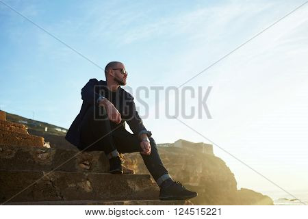 Young bearded hipster guy with trendy look is enjoying his recreation time during spring weekend abroad while is sitting on a rock cliff against sunset sky with copy space background for advertising