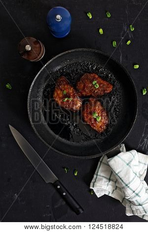 Korean style glazed pork chops on black background. Sticky citrus caramelized sauce with oranges and mint leaves. Salt, pepper, knife and green mint leaves near pan with chops.