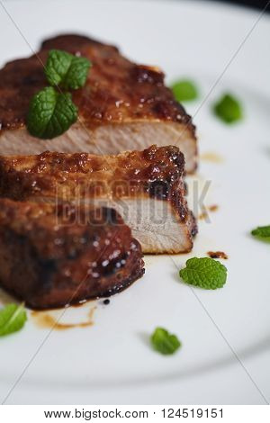 Korean style sliced glazed pork chops on white plate with sticky citrus caramelized sauce with oranges and mint leaves. Salt, pepper, knife and green mint leaves near pan with chops.