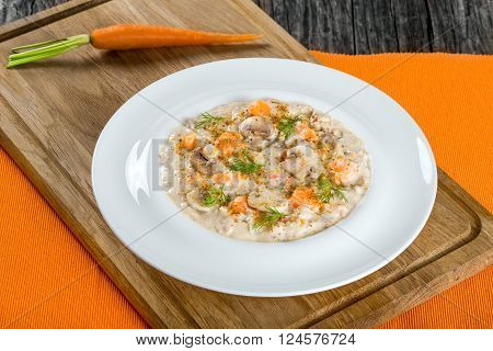 Delicious creamy cheese mushroom soup with spring carrots and spices decorated with dill in a white wide rim dish on a cutting board on an old rustic table horizontal close-up