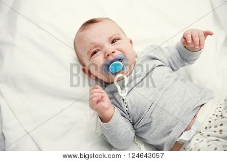 Crying baby with dummy lying on soft bed, close up