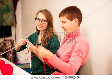 Women Knitting With Red Wool. Eldery Woman Transfering Her Knowledge Of Knitting On A Younger Woman