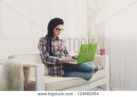 Young girl in protective computer glasses on a white sofa with laptop. Hipster freelancer woman works at home. Home office, freelance. Brunette with laptop in modern interior at white couch. Soft tone