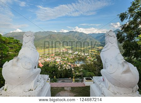 Back view of white lion statue at Wat Phra That Doi Kong Mu with aerial city view of Mae Hong Son against the mountain ranges, Thailand.