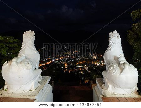 Back view of white lion statue at Wat Phra That Doi Kong Mu with aerial city night illuminated view of Mae Hong Son, Thailand