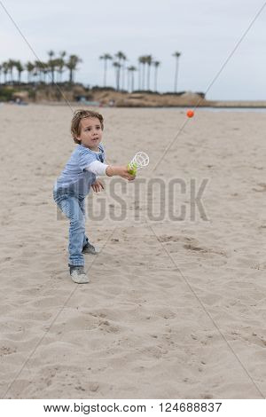 Lovely little boy on beach trying to catch orange ball with ball catcher. Palms on background