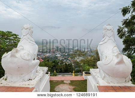 Back view of white lion statue at Wat Phra That Doi Kong Mu with aerial city view of Mae Hong Son in the mist, Thailand