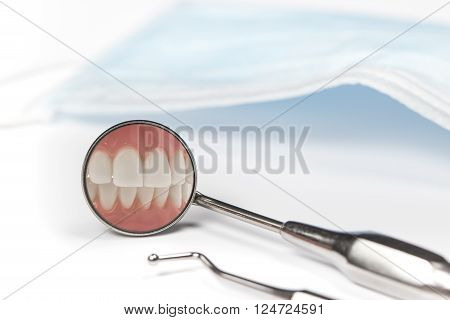 Dental mirror displays image of clenched front teeth beside metal pick and mask on white table