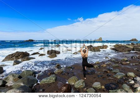Summer yoga session on beautiful Playa de Roque de Las Bodegas with giant rock and high waves - tropical Tenerife island, Canary in Spain. Vriksha-asana, tree pose