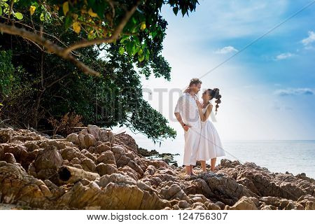 Elegant smiling young bride and groom walking on the beach, kissing and having fun, wedding ceremony near the rocks and ocean